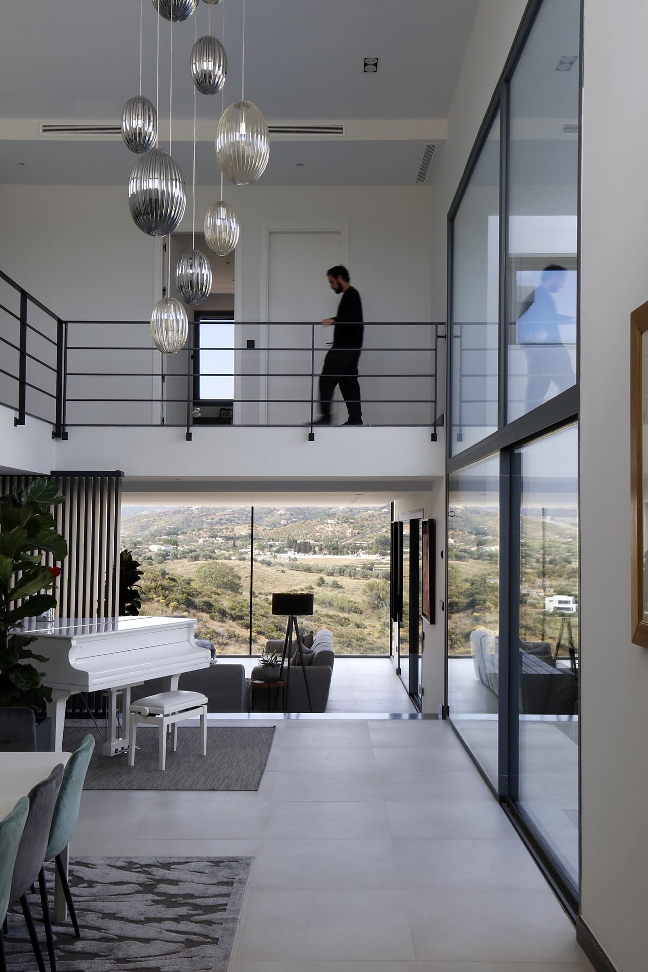 Interior de doble altura con pasarela metálica, lámparas colgantes esféricas y vista al paisaje exterior