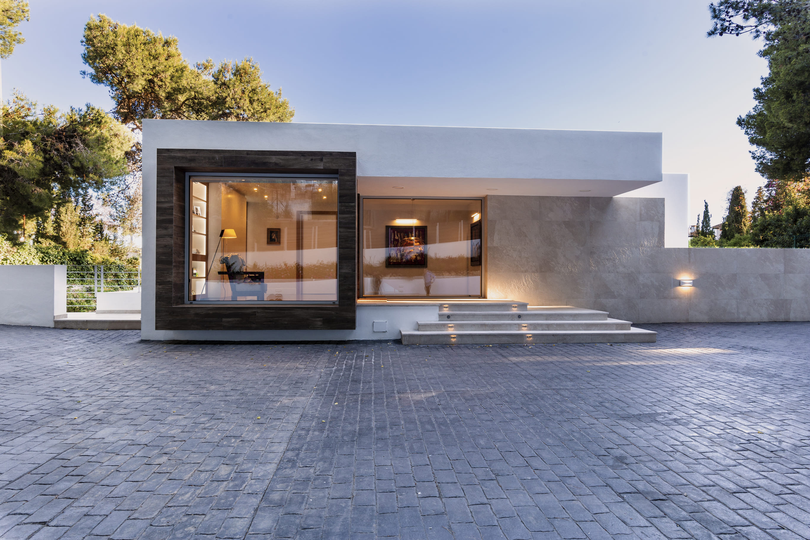 Vista diurna de la entrada pavimentada de la Vivienda P∩C con volúmenes blancos y entorno de pinos