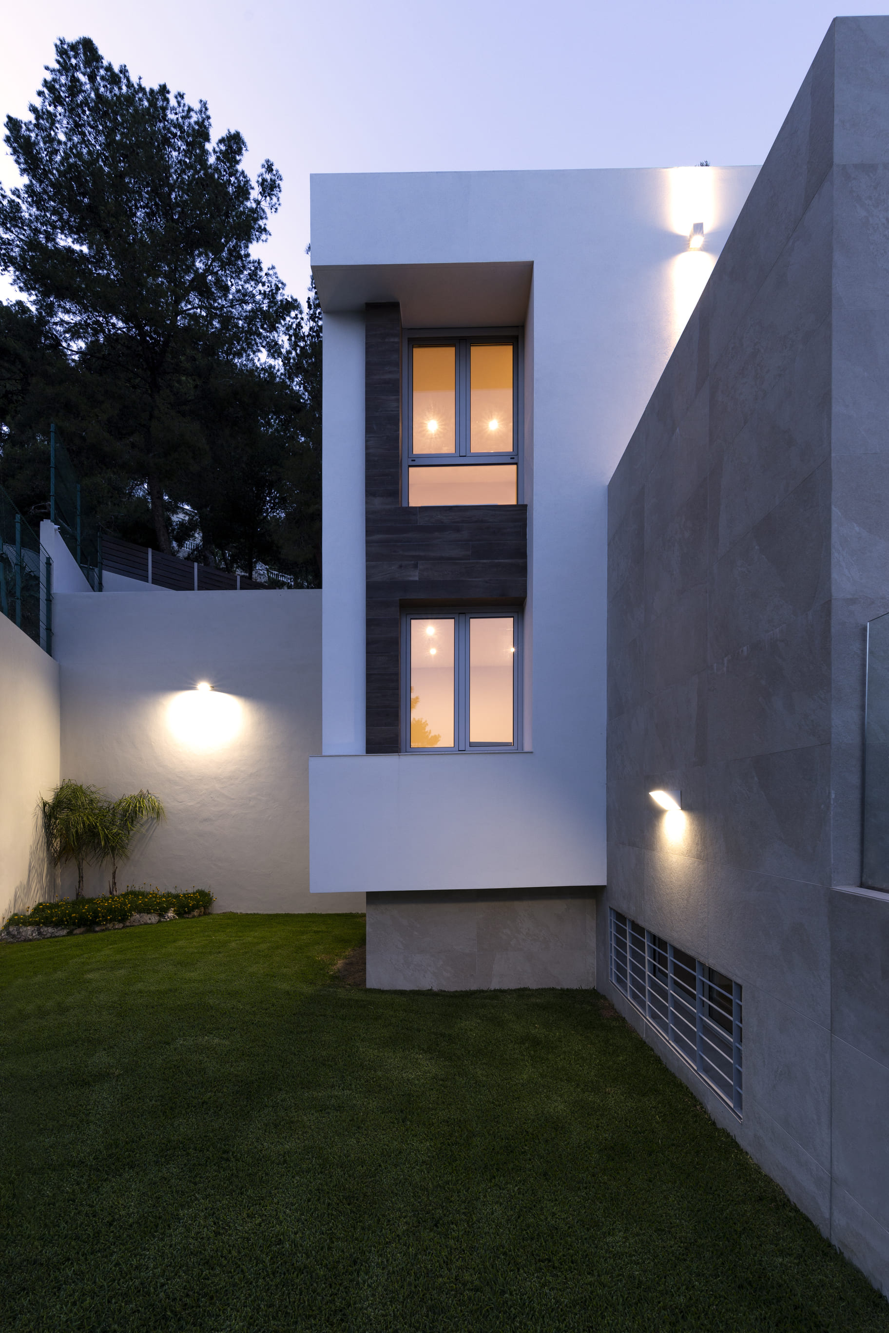 Vista lateral de la Vivienda P∩C con fachada blanca y ventana vertical iluminada por la noche